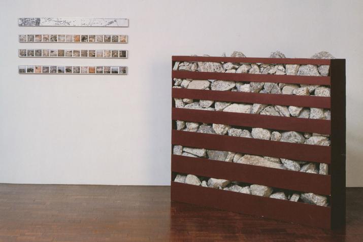 A metal box filled with rocks and horizontal slots cut in the box on the floor and horizontal images on the wall.