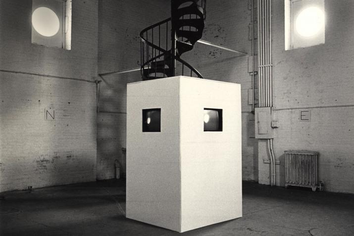 a white box with two screens in embedded inside sits in the center of a white brick room with a spiral staircase at the back and two high windows.
