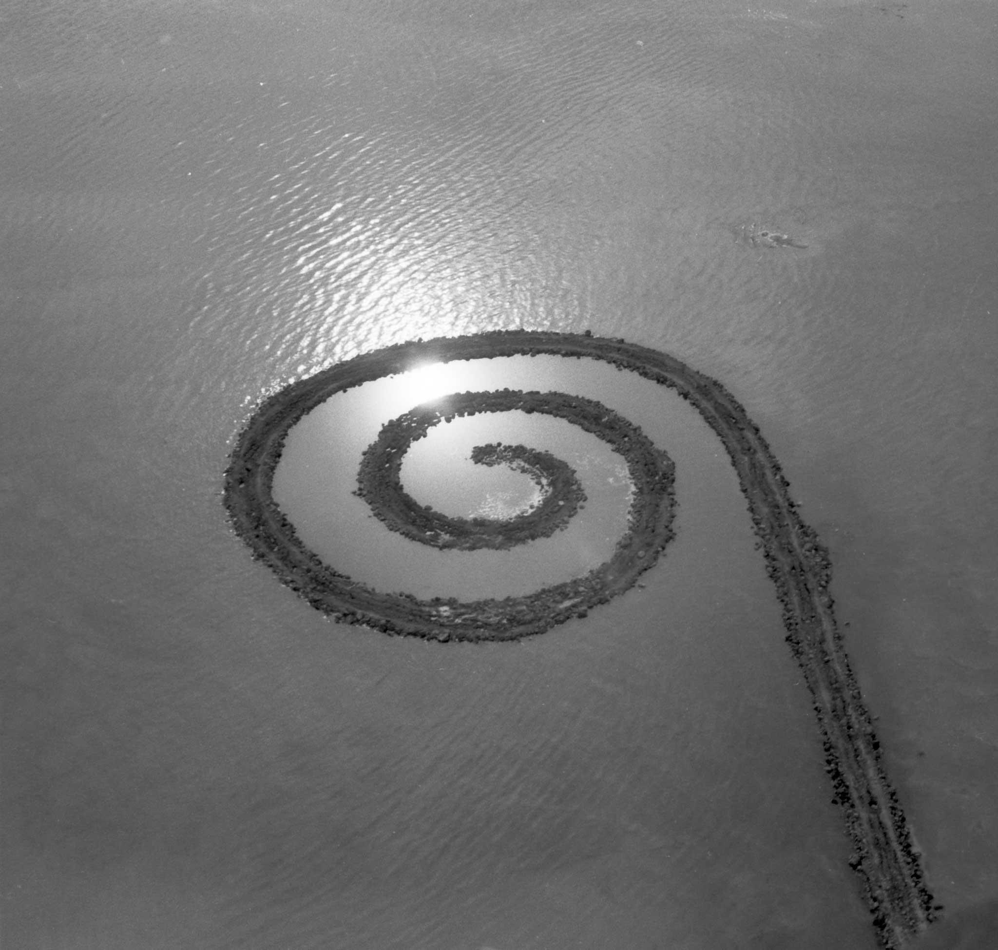 black and white, aerial image or an earthen jetty with a spiral termination extending into a body of water 