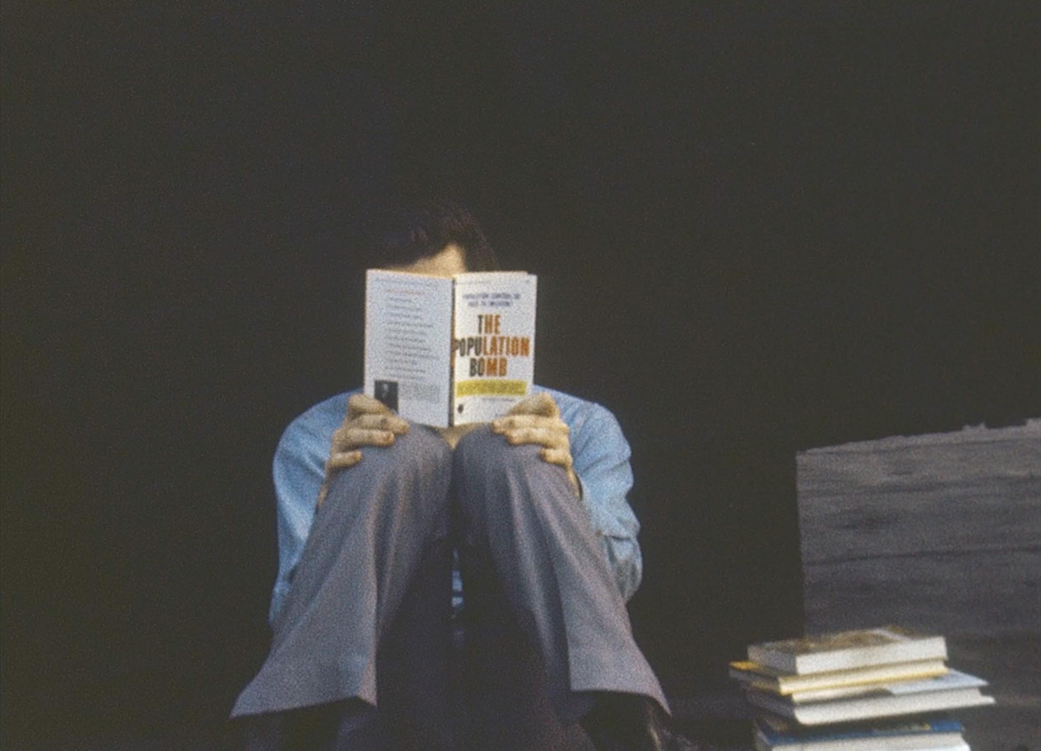 person sitting on the ground on a rooftop reading a book with knees together and book obscuring their face