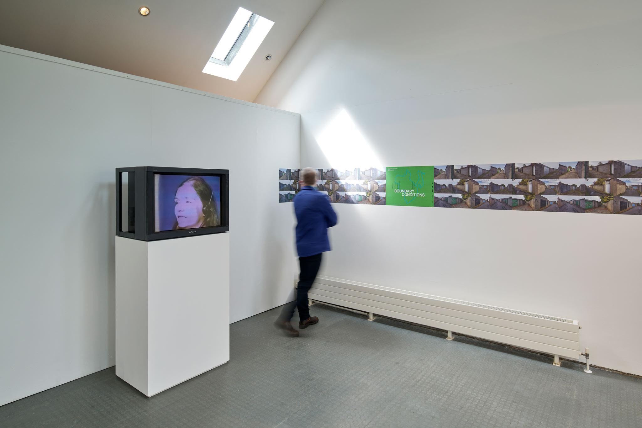 a person standing in a gallery space with a monitor on the left and a poster on the right