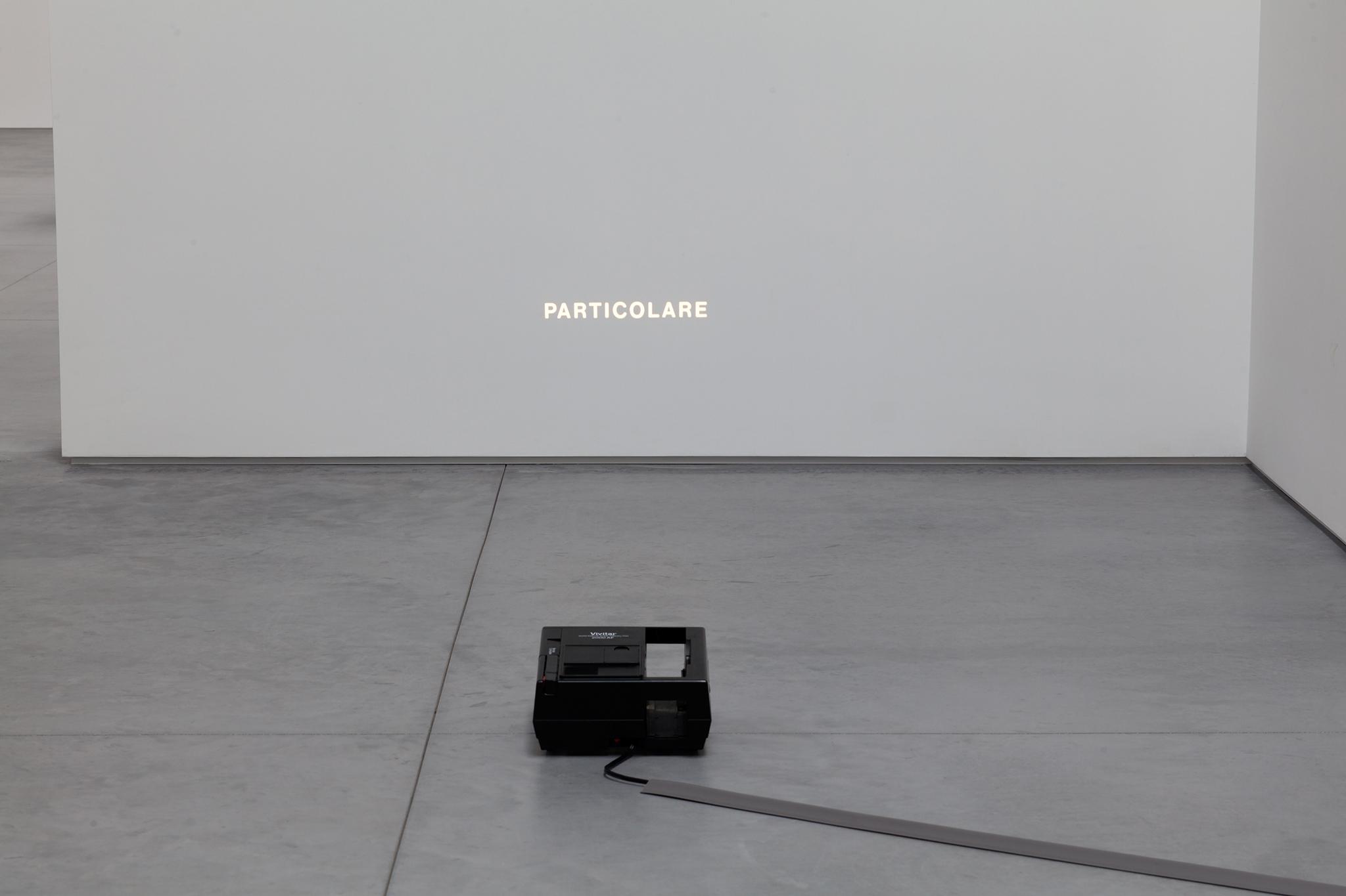 a slide projector sitting the floor projecting the word particolare onto a white wall