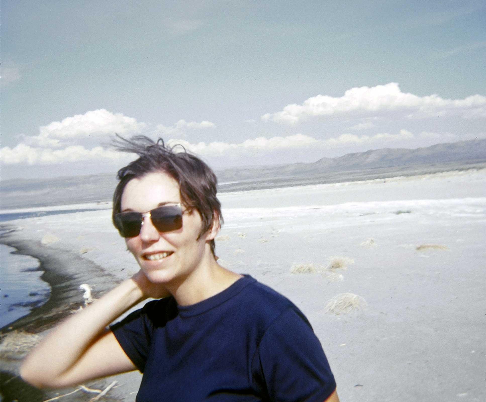 Nancy Holt standing by shore of lake with sunglasses on and short brown hair.