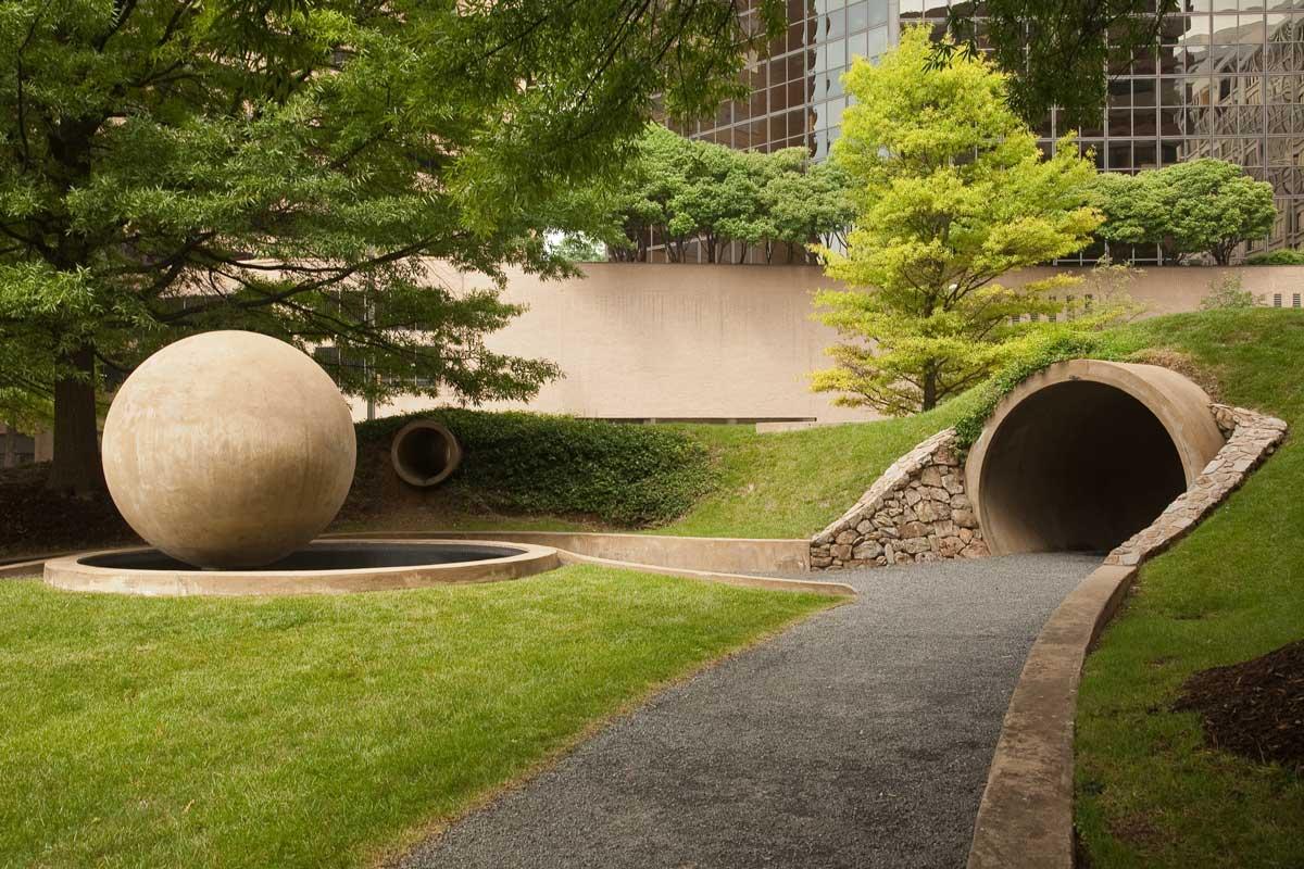 Gravel path next to a green grassy lawn with a concrete sphere sculpture and a round concrete tunnel.  Trees in the background.