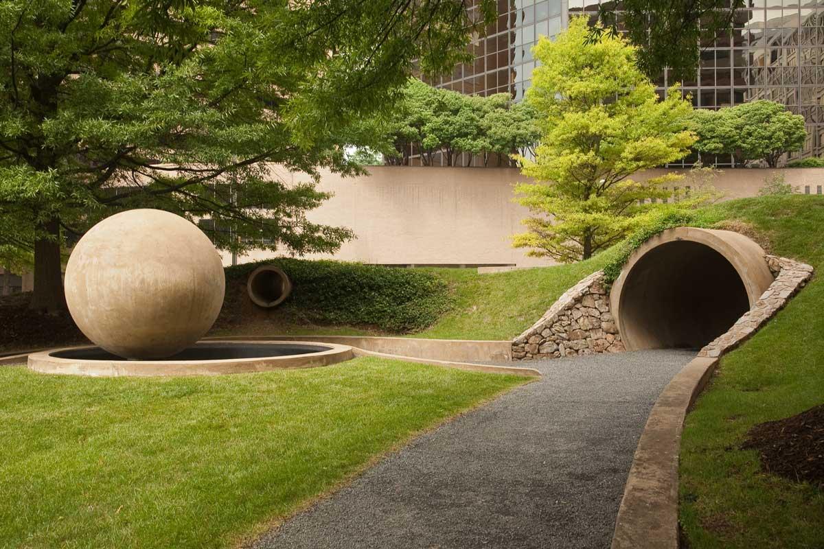Gravel path next to a green grassy lawn with a concrete sphere sculpture and a round concrete tunnel. Trees in the background.