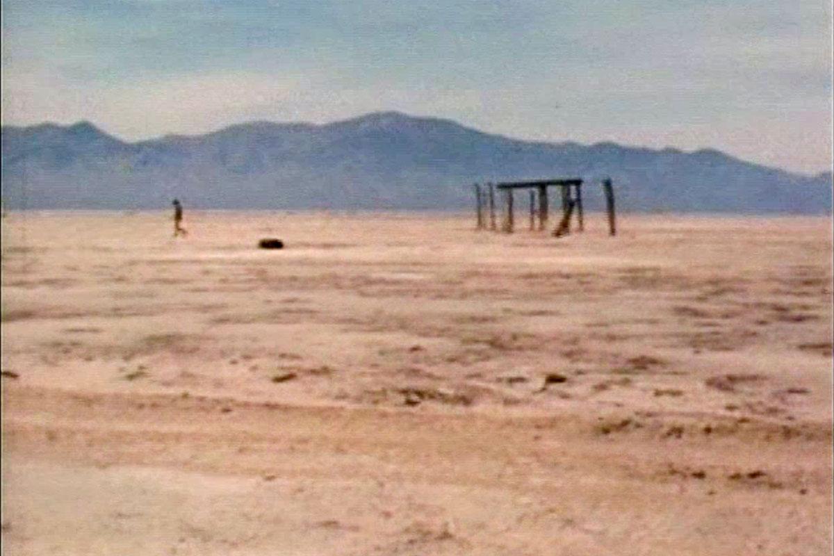 Desert in the foreground with blue mountains in the background.  In the mid-ground is a dilapidated wood structure and a figure.