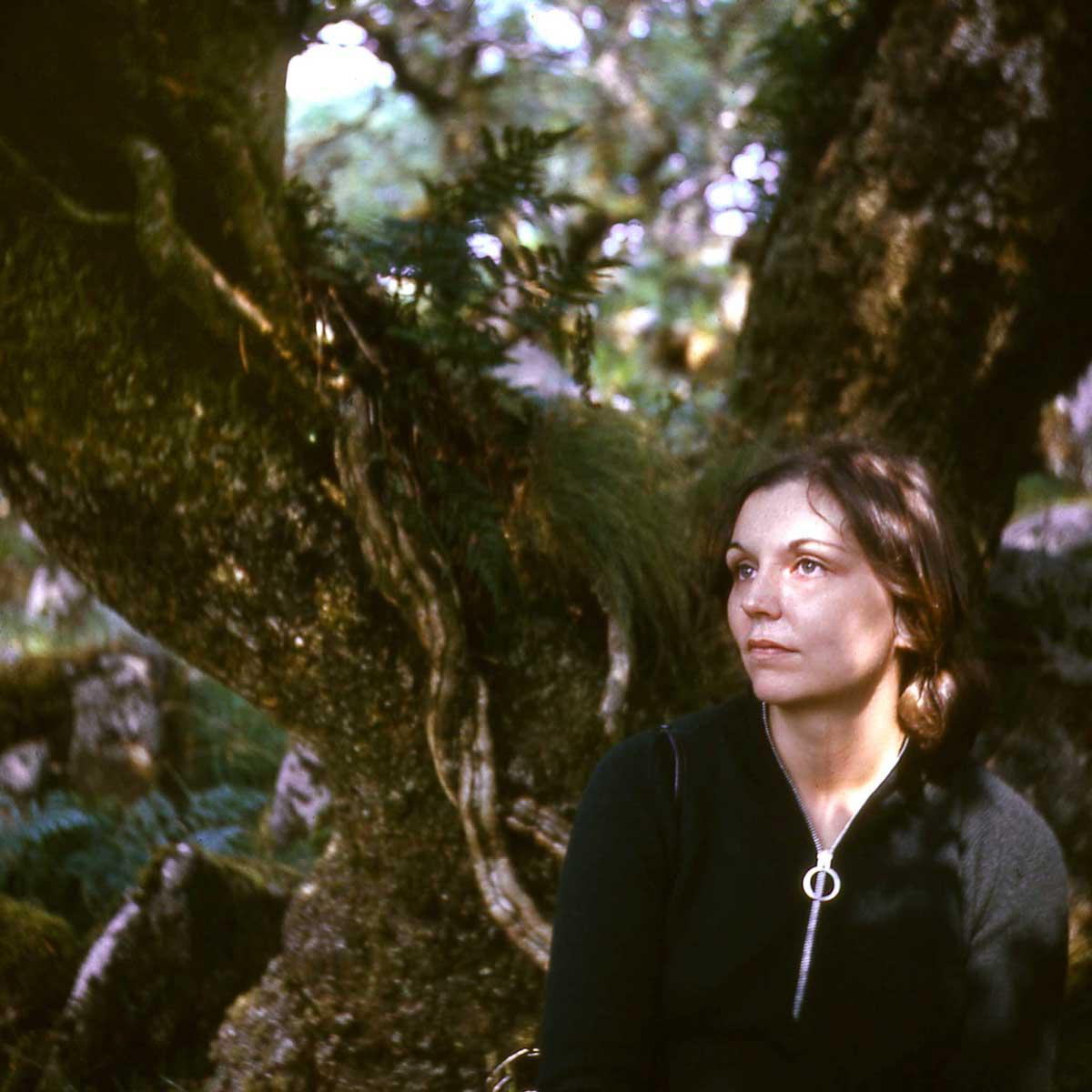 A woman leaning against a mossy tree with dappled light