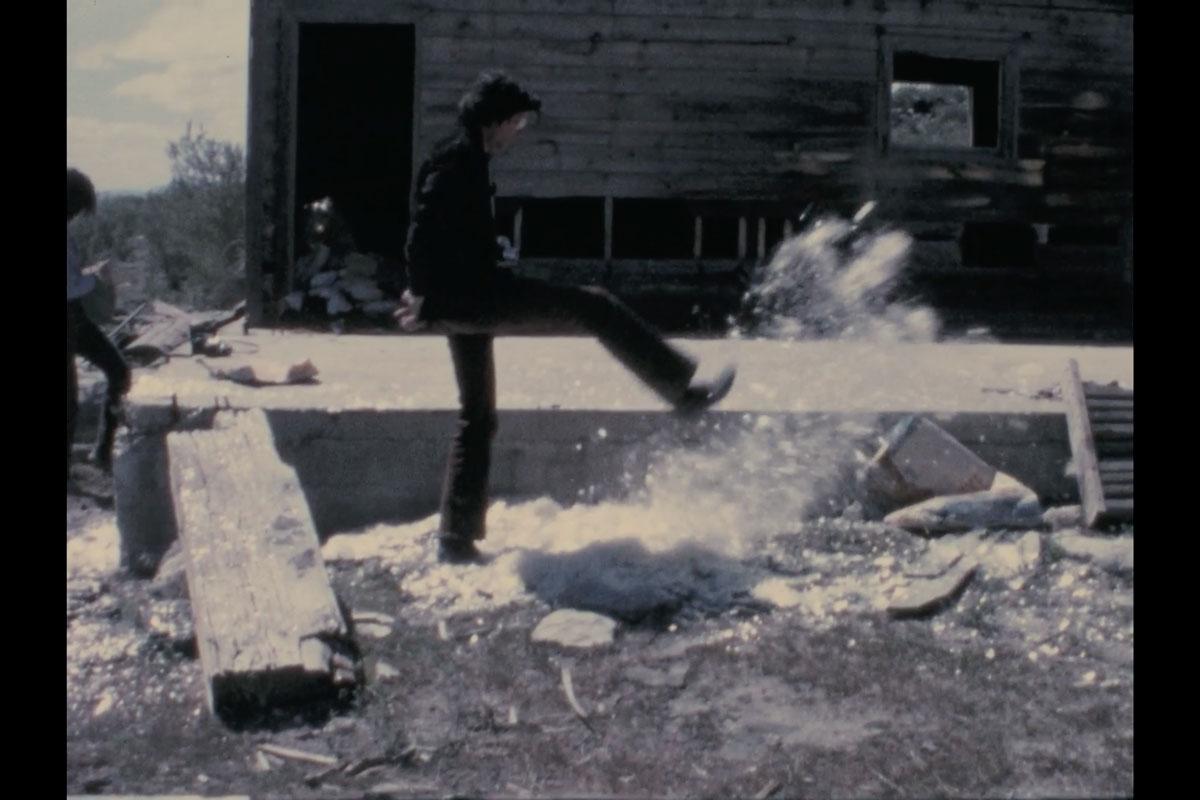 a man kicking a pile of mica and the mica is flying up in the air in front of a small wood cabin