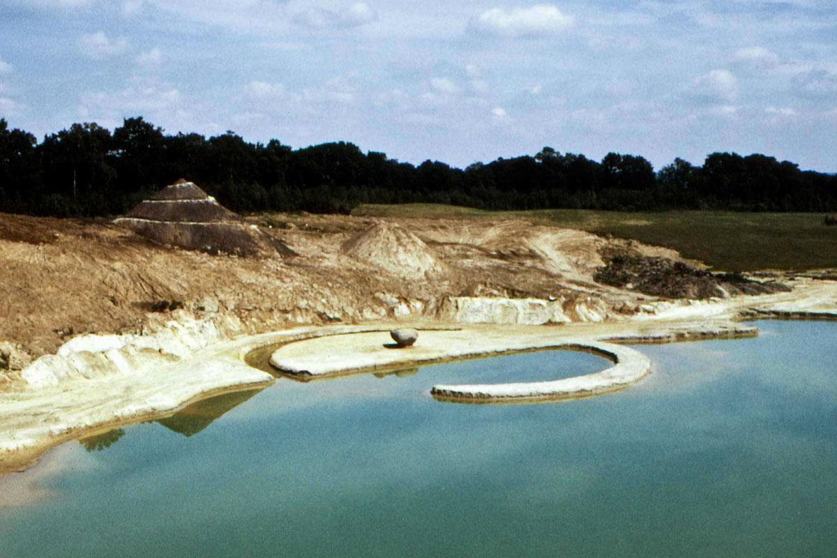 a sand quary with a jetty, canal, and a spiraling hill of earth and trees in the background