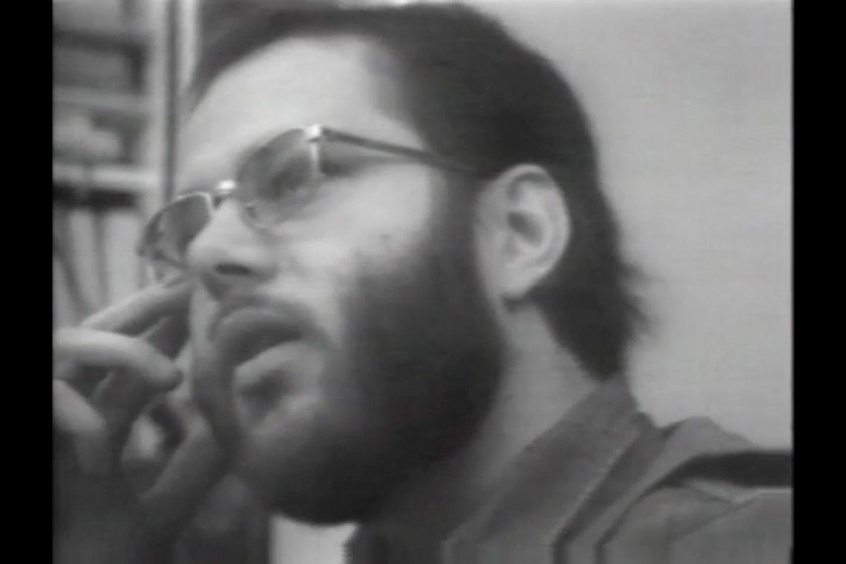 Black and white image of a man with a beard and glasses talking looking to the left.