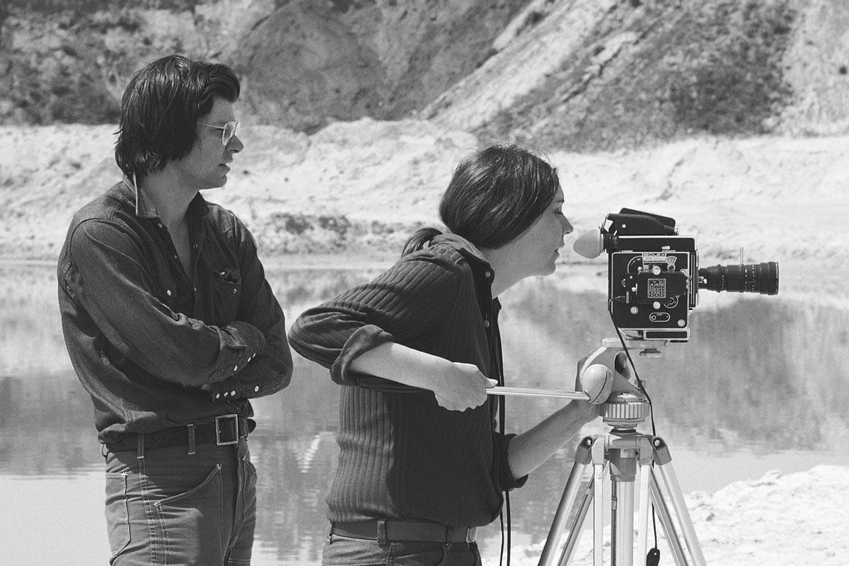 black and white image of two figures. One stands behind and one looks through the viewfinder of a Bolex film camera on a tripod