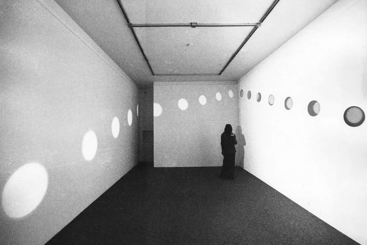 A figure standing in a white walled room. A diagonal line of mirrors in a white walled room. Light is reflecting off the mirrors, creating orbs of light on the other two walls