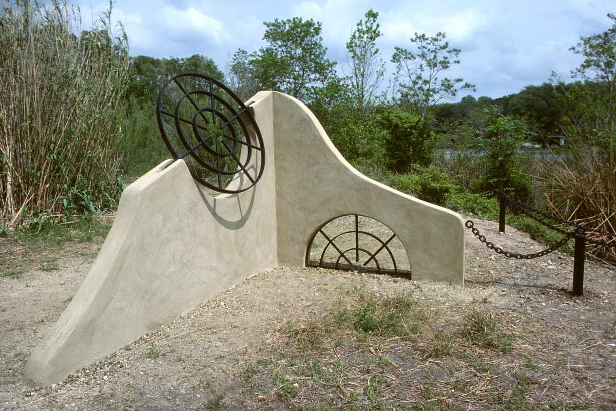 a concrete and metal sculpture of an undulating wall and circular steel inset into the wall