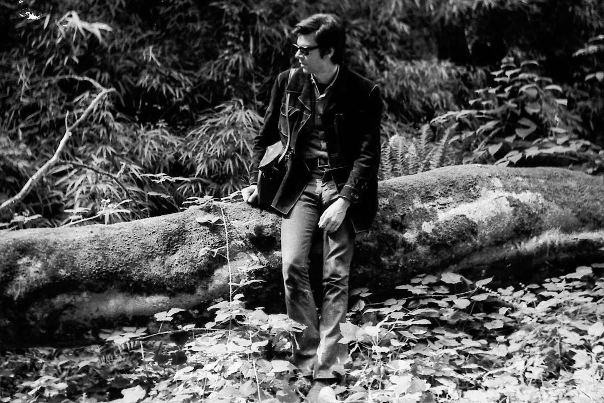 black and white image of a person looking to the left while sitting on a large log in the forest 