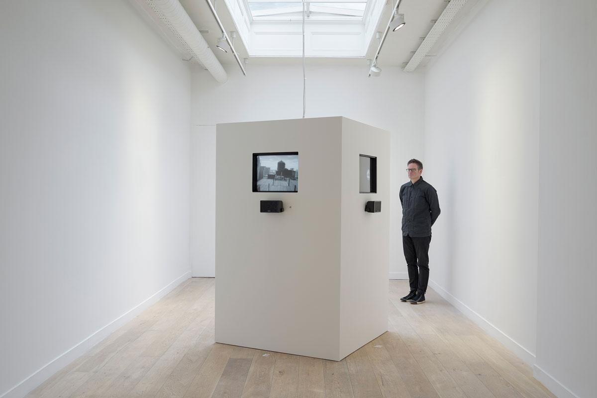 white room with a large white box in the center containing television monitors playing black and white video. One figure standing watching