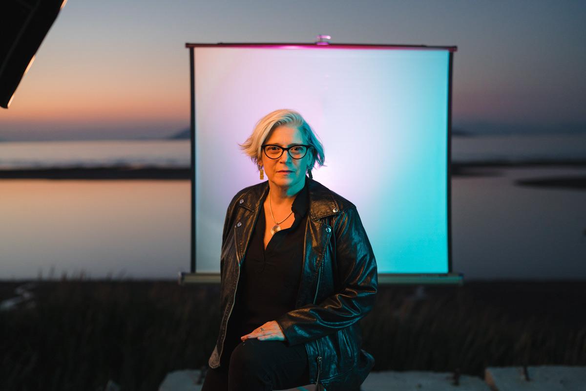 a figure wearing all black and medium length gray hair is seated and faces the camera with a lake at sunset in the background