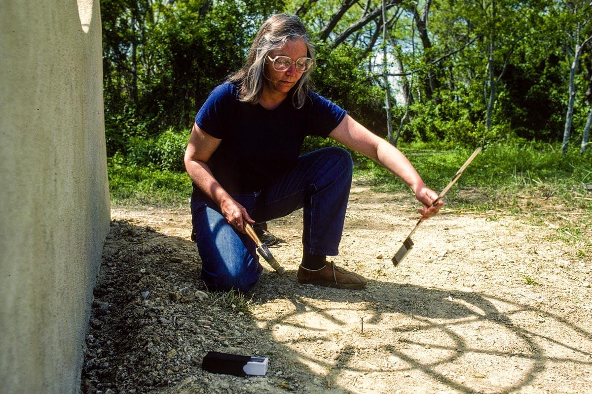 person kneeling beside a sculptural wall, and using tools to mark the alignment of the shadow of the sculpture