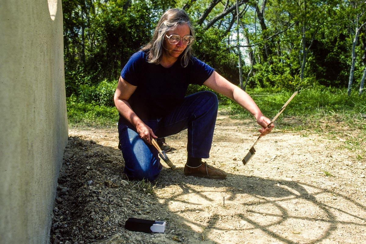 person kneeling beside a sculptural wall, and using tools to mark the alignment of the shadow of the sculpture 