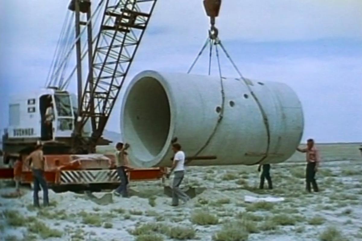a crane lifts a large tunnel into place with guidance from five figures on the ground