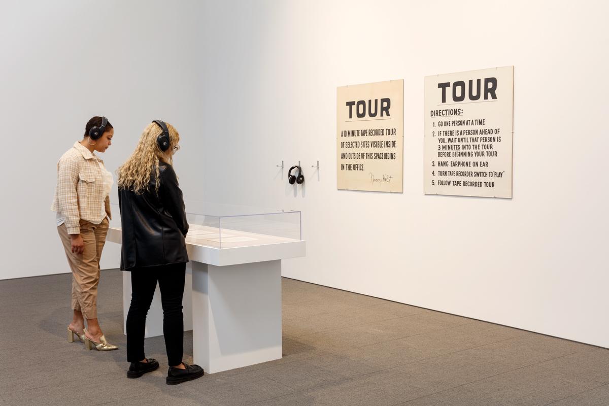 two people standing at a vitrine with headphones on for the Tour of the John Weber Gallery by Nancy Holt