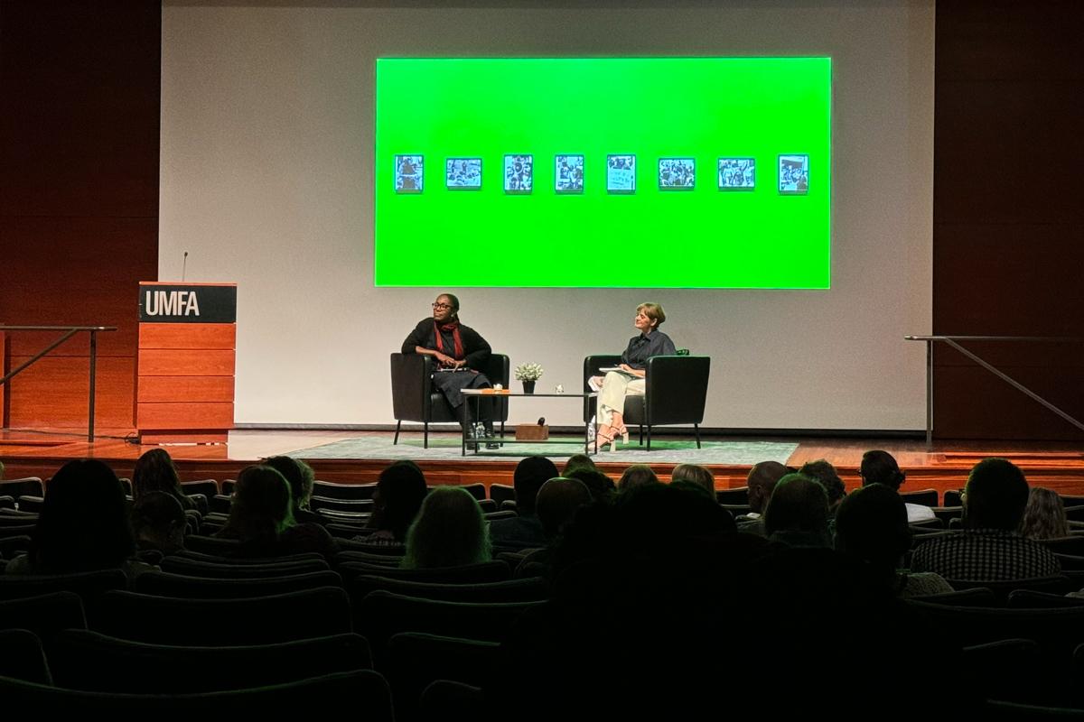 two people sitting on a stage with a projected green image behind them and with a crowd in the foreground