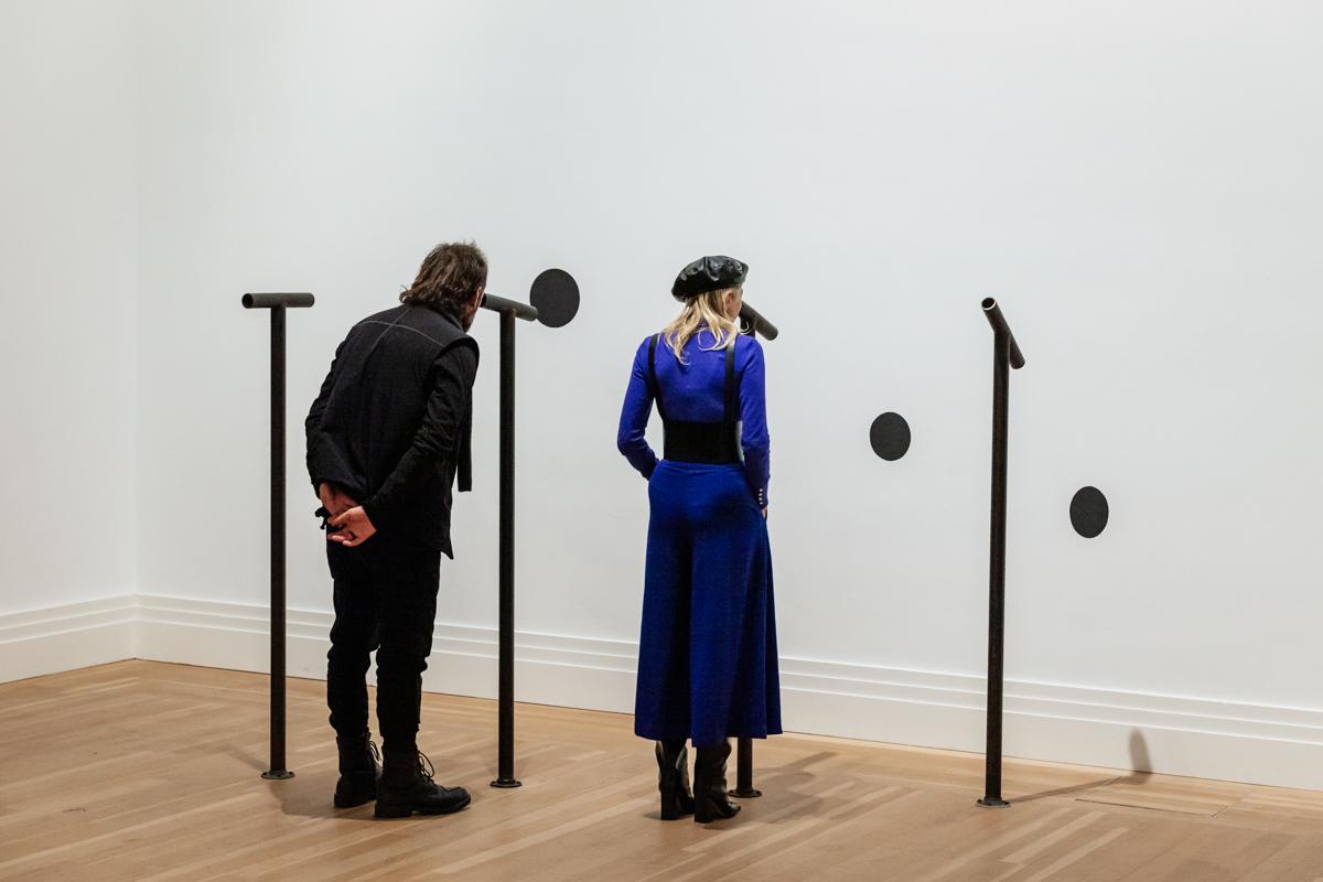 two people looking through sculptural seeing devices at black circular shapes painted on a white wall