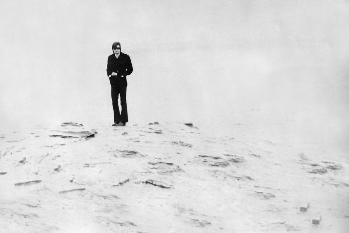 black and white image of man standing on shoreline wearing all black. The foreground and background are very light.
