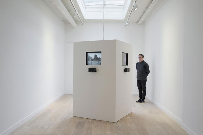 white room with a large white box in the center containing television monitors playing black and white video. One figure standing watching