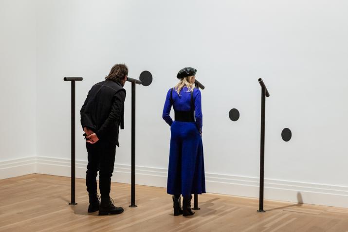 two people looking through sculptural seeing devices at black circular shapes painted on a white wall