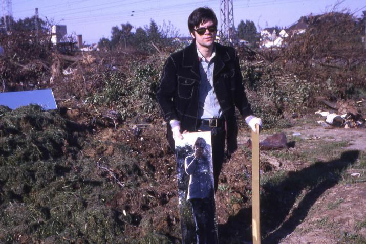 A man standing next to a compost heap holding a mirror beside his leg, which reflects a woman holding a camera