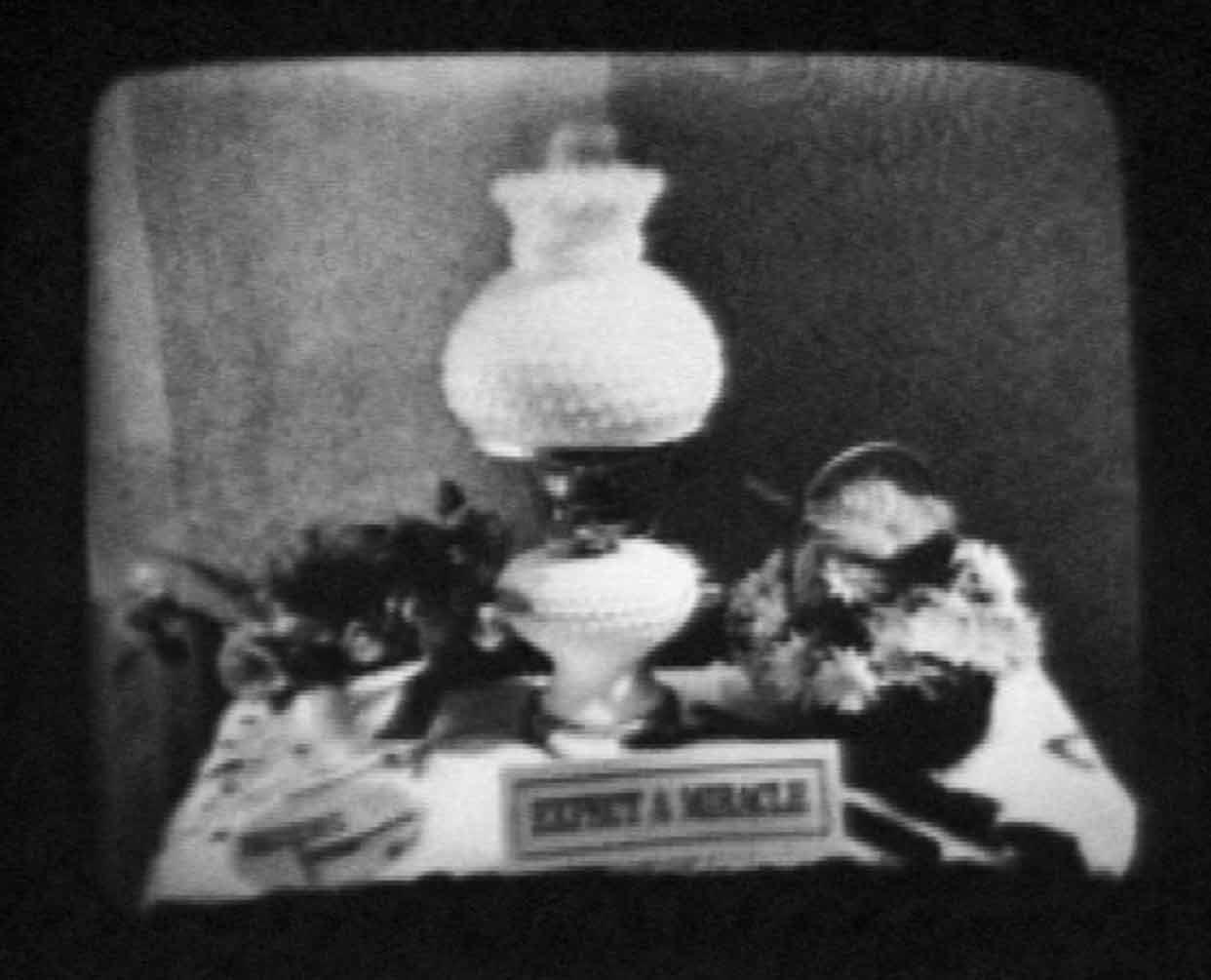 black and white image of a lamp on a messy desk and a small sign that reads "expect a miracle"