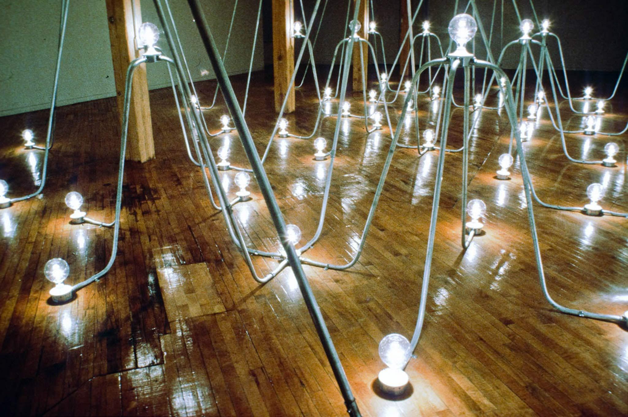 Room with no windows lit with numerous lightbulbs that are connected via an arching rhythmic armature of steel conduit.