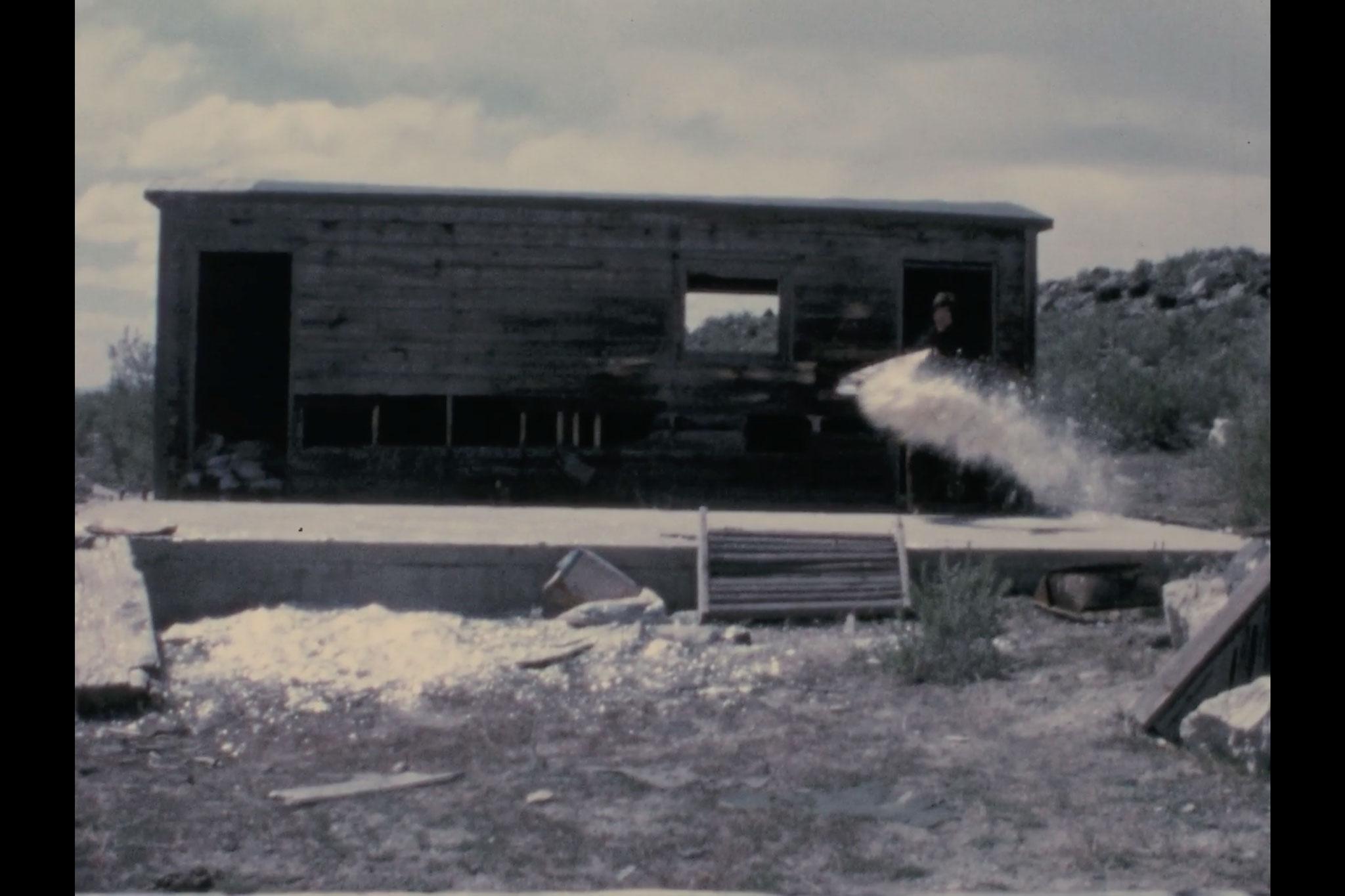 A small cabin with a man standing on the front porch throwing the contents of a bag of mica into the air