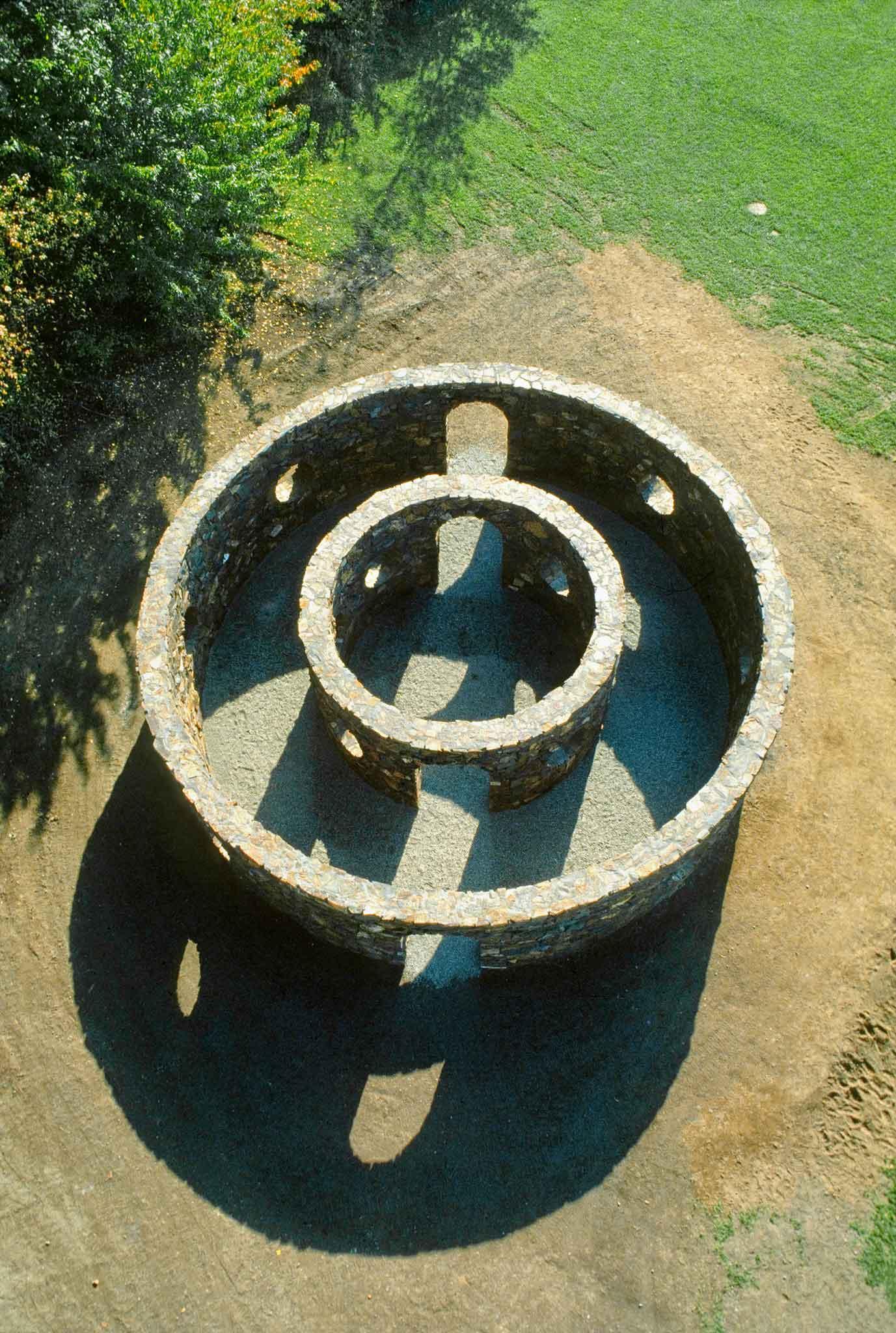 An aerial view of two concentric circular stone walls with the forest surrounding on the left and grass on the right.