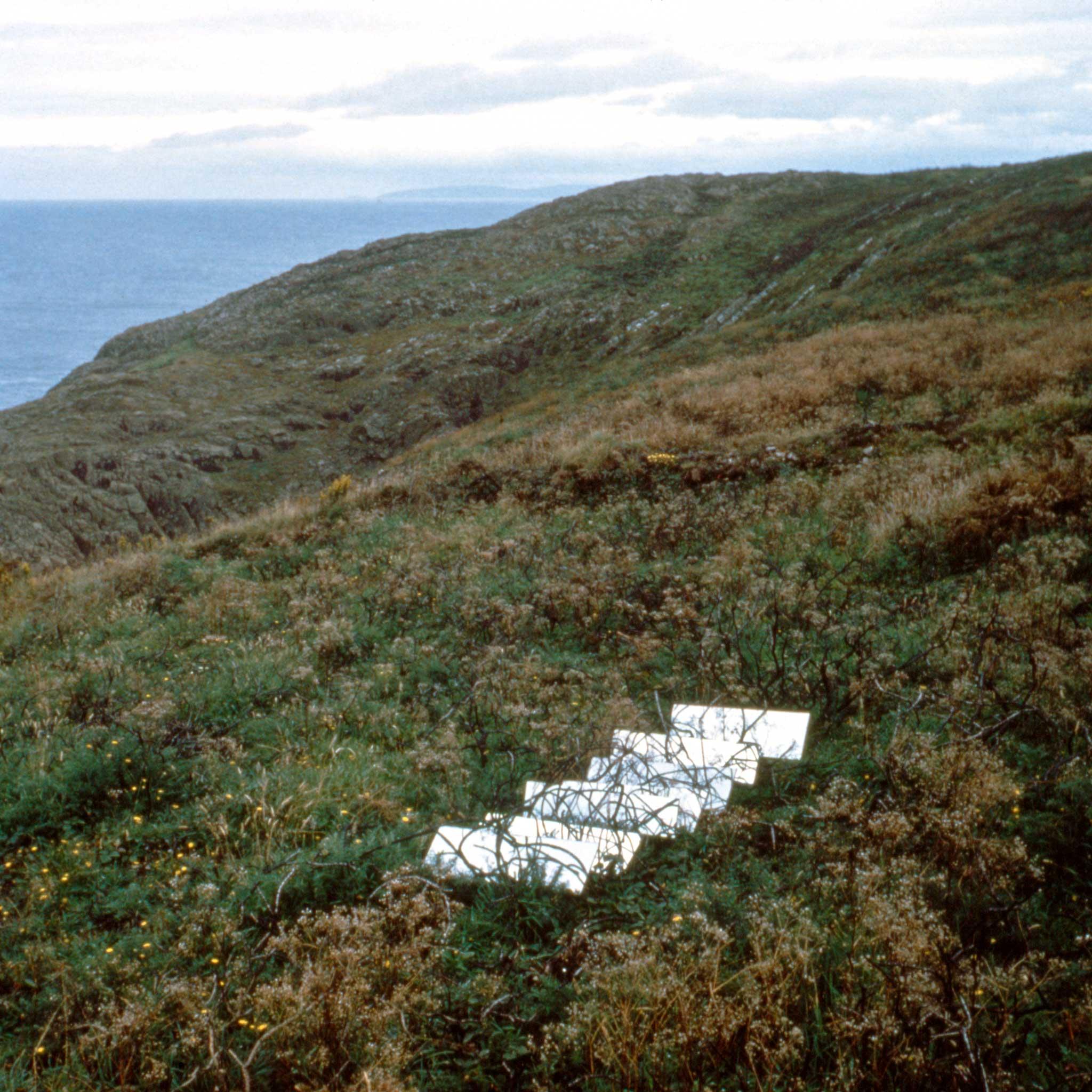 grassy hilly landscape with ocean in background with seven mirrors placed on the ground.