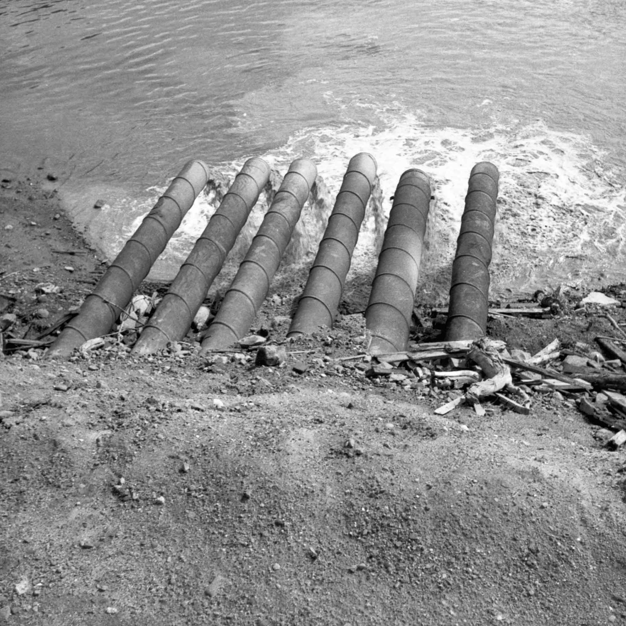 black and white image of six metal tubes jutting out of an earthen bank dumping water into a body of water. top down view