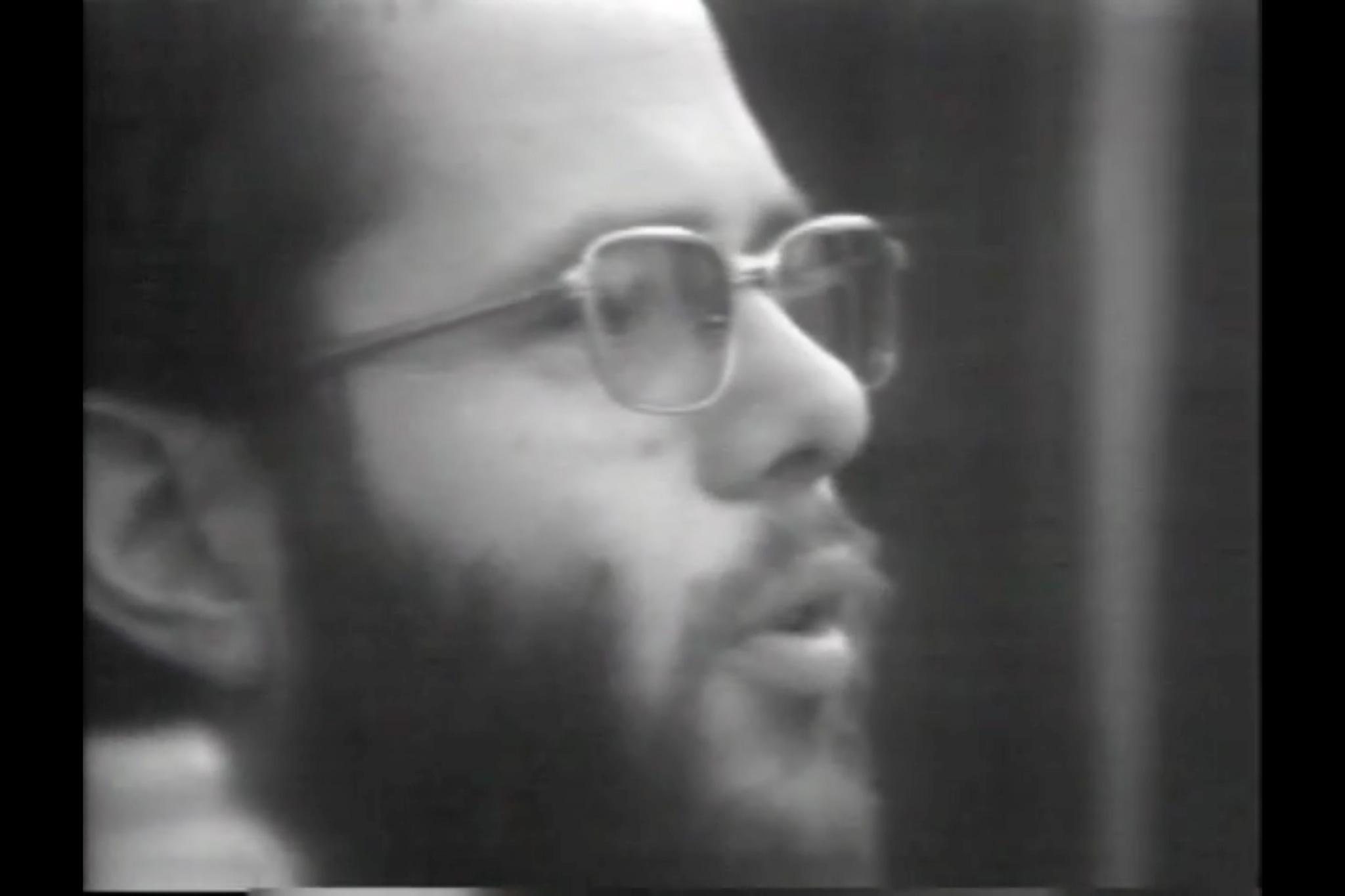 Black and white image of a man with a beard and glasses talking looking to the right.