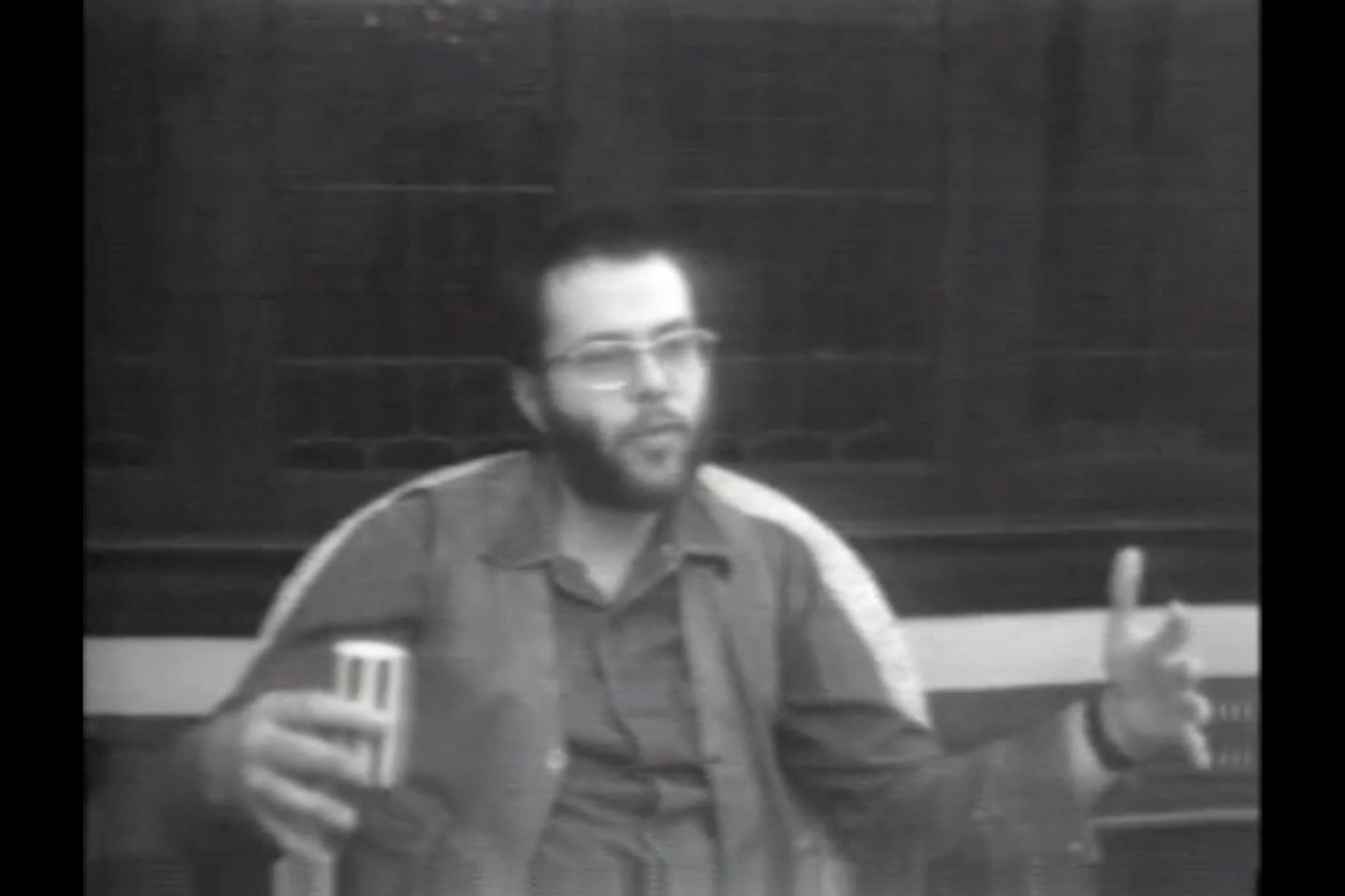 Black and white image of a man with a beard and glasses talking and gesturing.