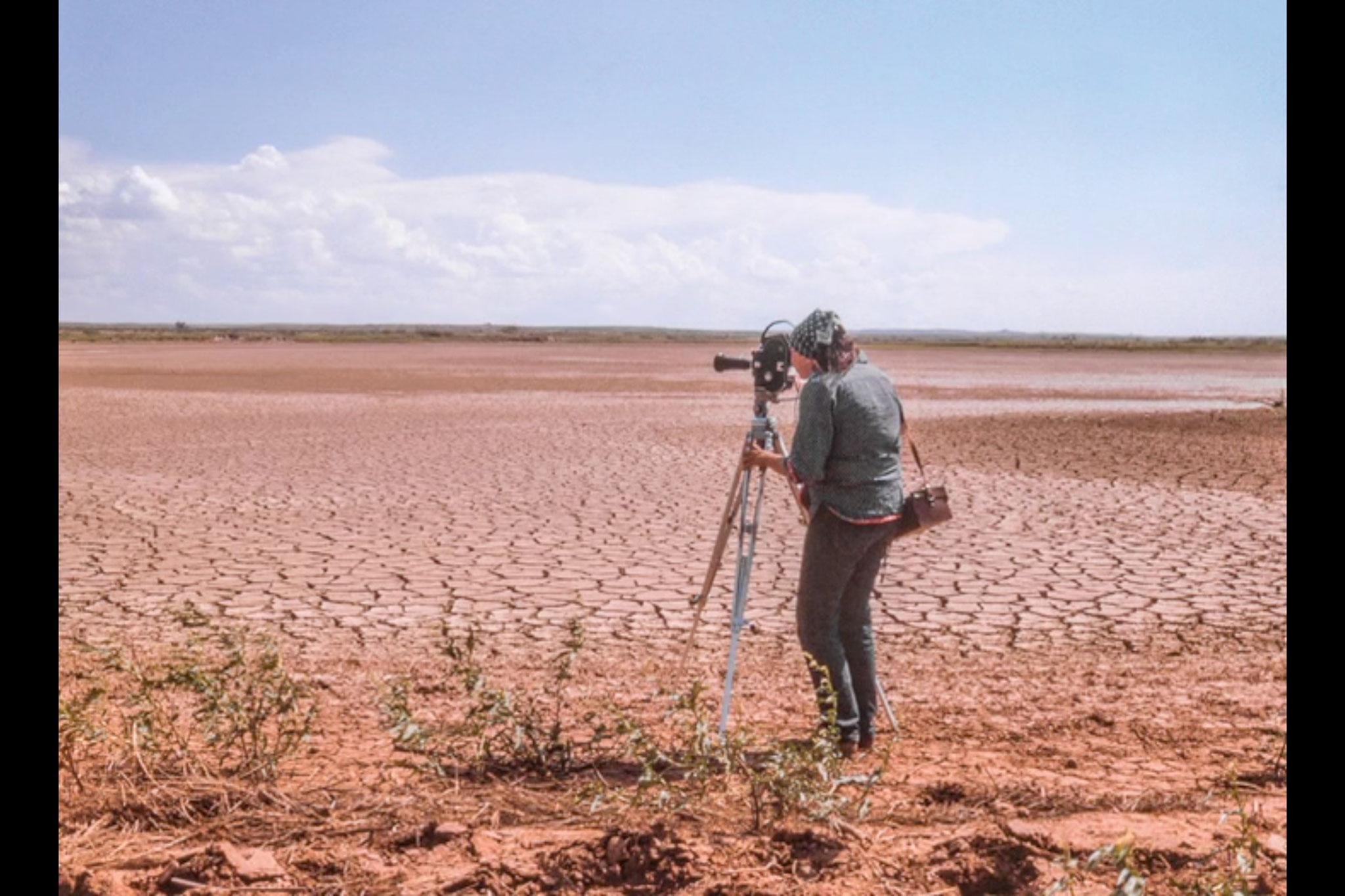 A woman standing next to a film camera and tripod on the edge of a dry lakebed.