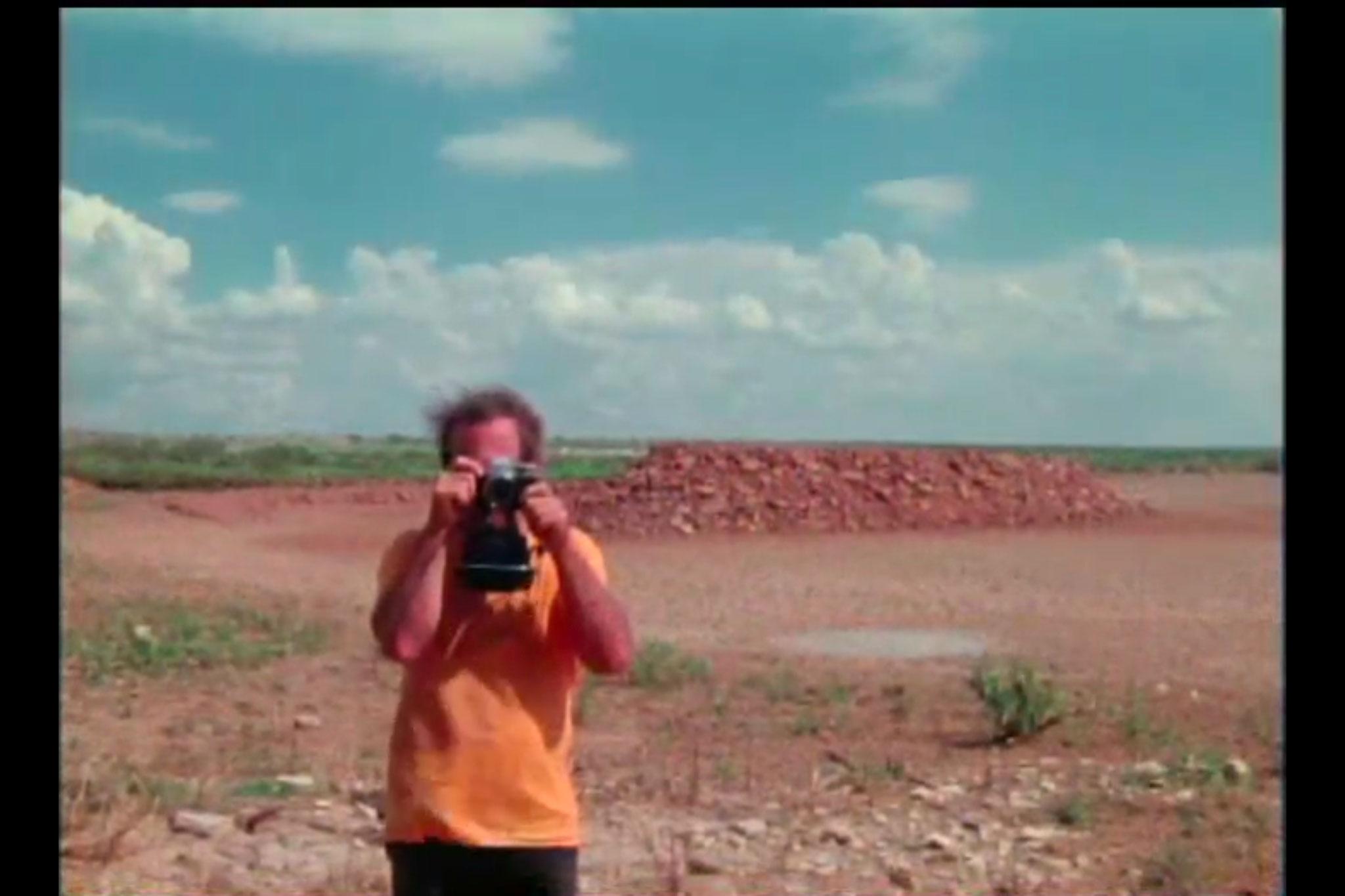 A young man taking a picture toward the camera with a dry lakebed in the background.