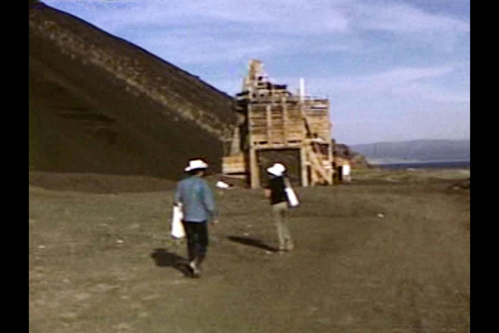 two figures walking toward an industrial building with a slope of gravel in the background.