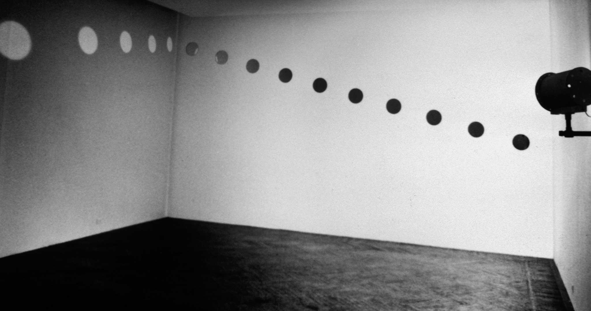 A diagonal line of mirrors and circular orbs of light reflected on two white walls of a room with no windows