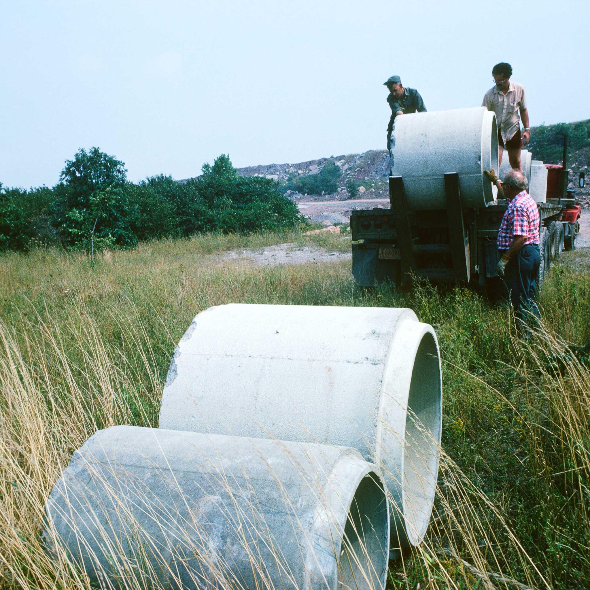 Two large concrete tubes in a field and a third on a front loader machine with three figures around it.