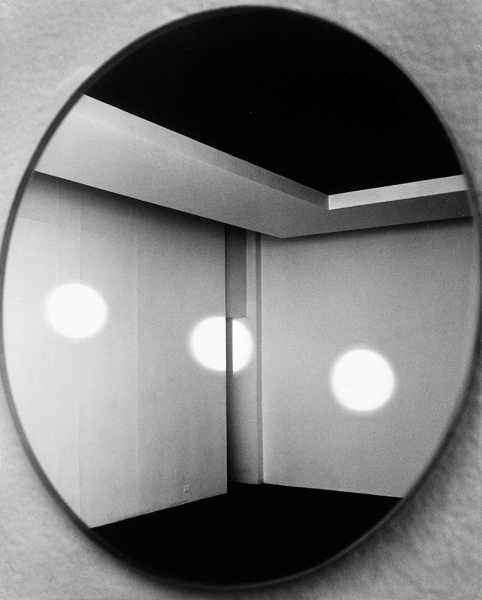 a closeup of a circular mirror, which reflects the back corner of a room with white walls and no windows
