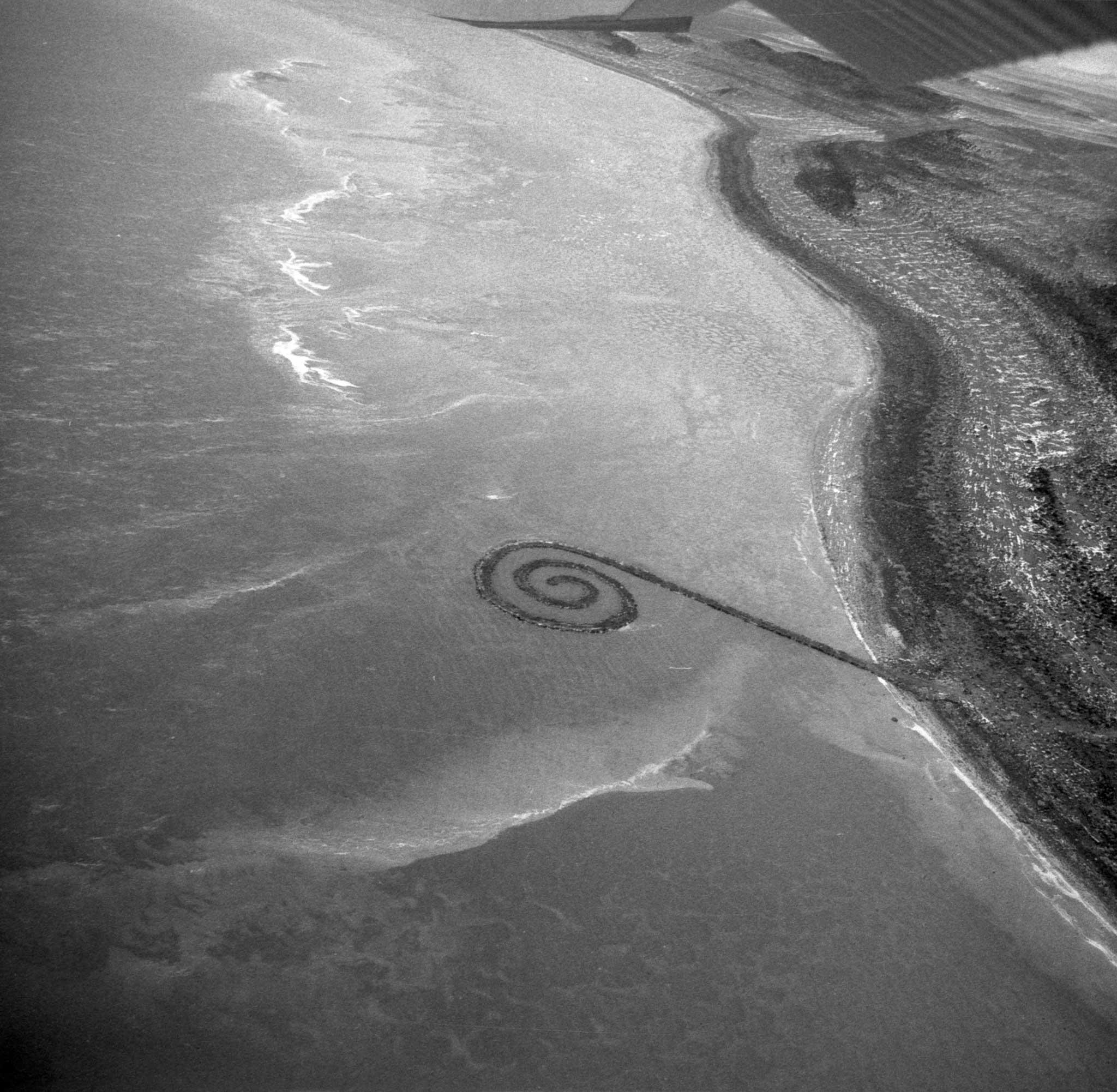 black and white, aerial image or an earthen jetty with a spiral termination extending out from the shore into a body of water 