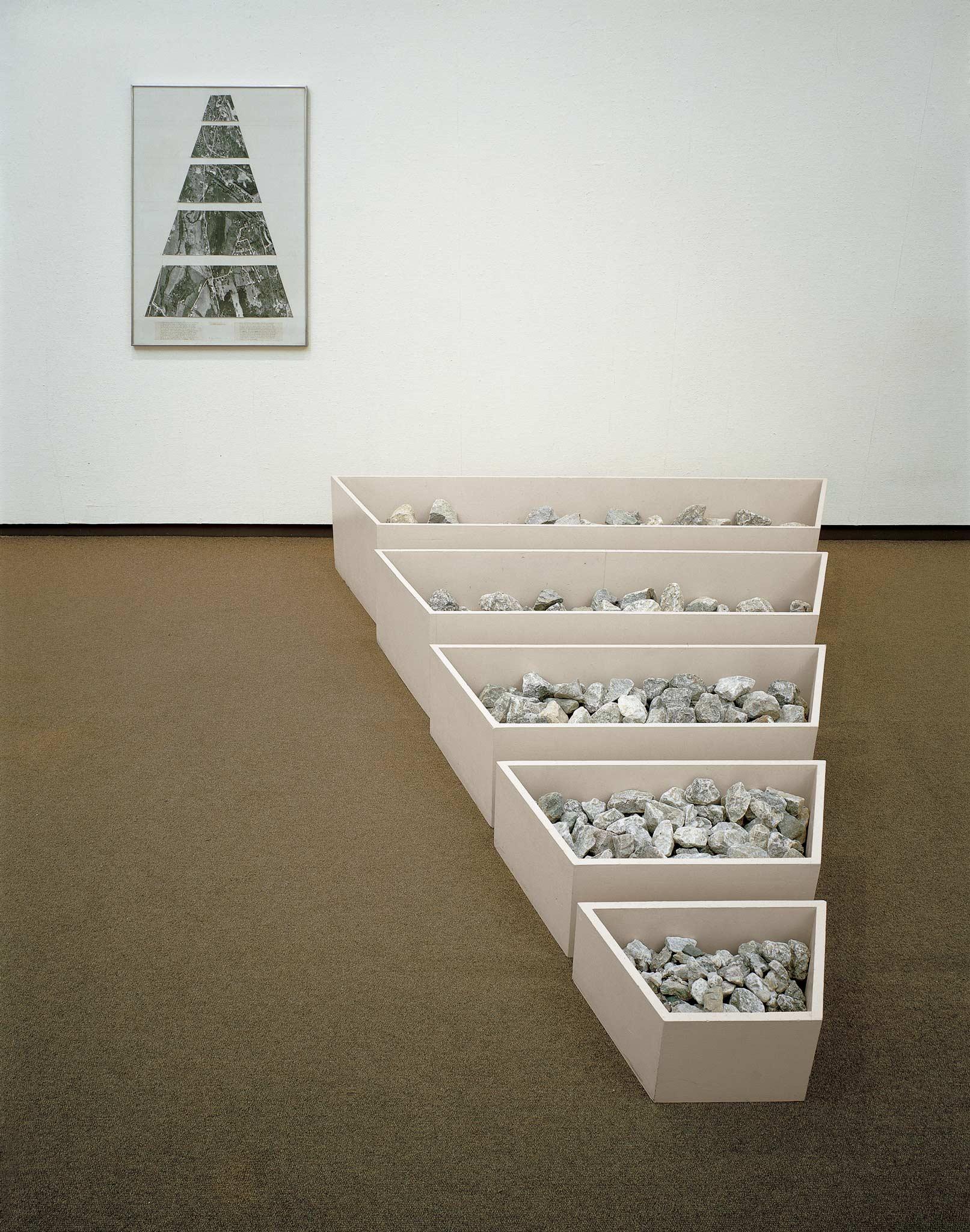Five low bins full of rocks arranged on the floor from largest to smallest. On the wall is a framed collage of a map that shows the same shapes.