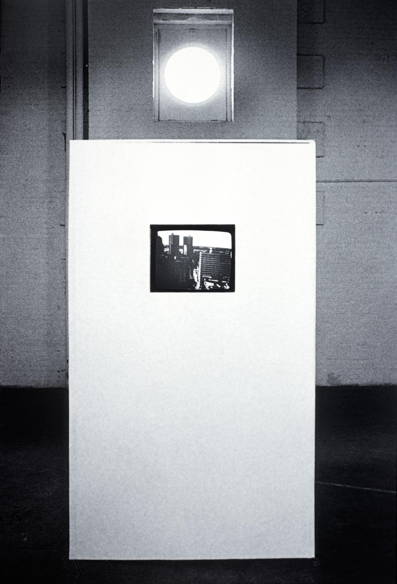 a white box with a screens embedded inside sits in the center of a white brick room with a high circular window in the background.