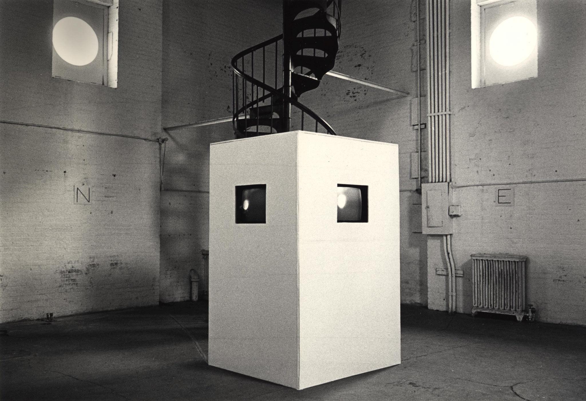 a white box with two screens in embedded inside sits in the center of a white brick room with a spiral staircase at the back and two high windows.