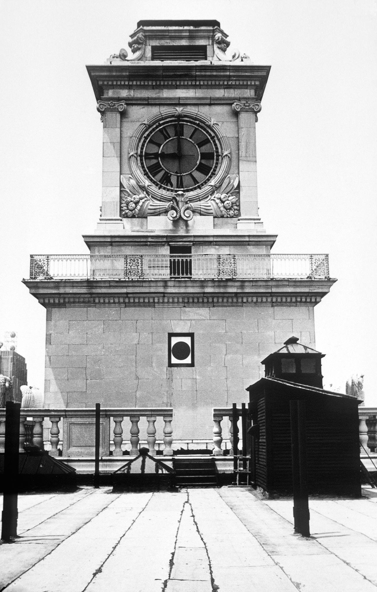 A stone clocktower with a small circular window below the center of the clock face. Black and white.