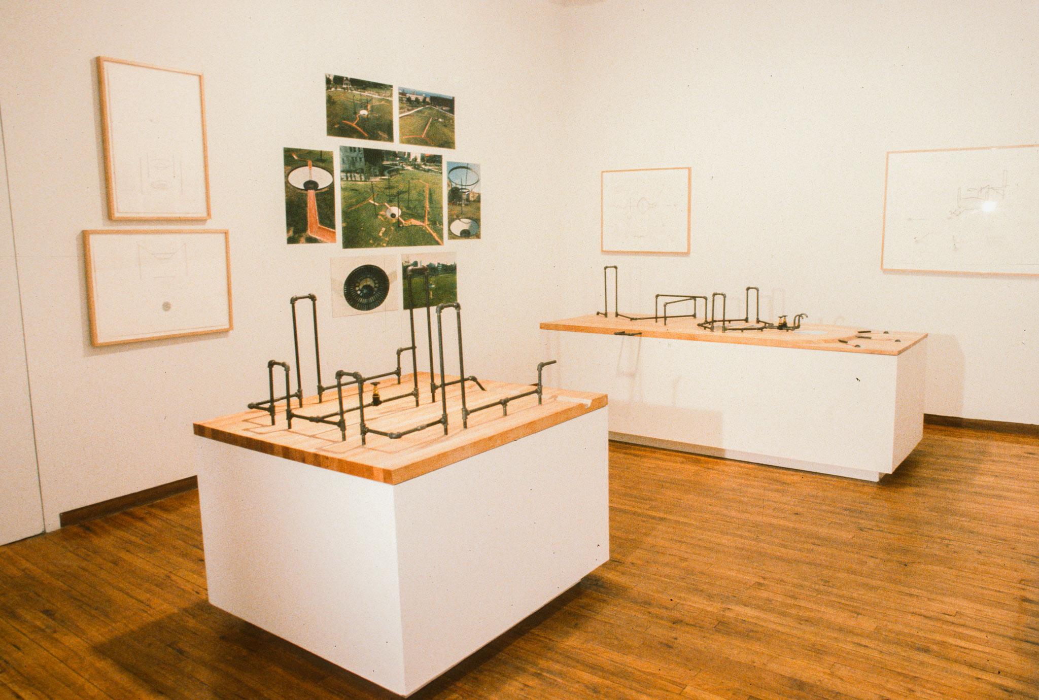 a gallery with drawings and photographs on the wall and two small metal pipe models on pedestals  